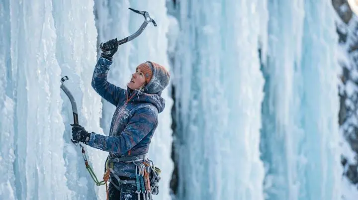 A climber swinging an ice tool into a frozen waterfall, focused and determined, surrounded by blue ice formations