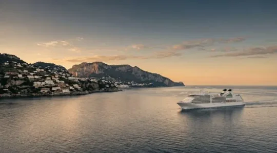 Wide panoramic view of a cruise ship sailing at golden hour past a Mediterranean coastal town, symbolizing a multi-country voyage
