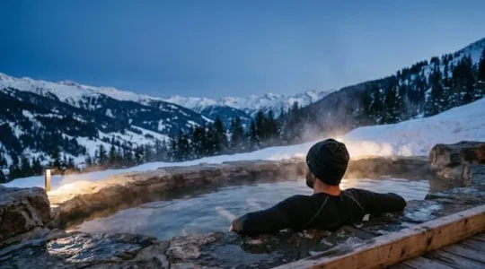 A skier relaxing in an outdoor mountain thermal pool surrounded by snow at dusk, symbolizing post-ski muscle recovery