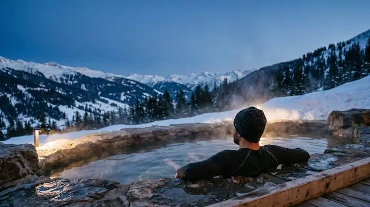 A skier relaxing in an outdoor mountain thermal pool surrounded by snow at dusk, symbolizing post-ski muscle recovery