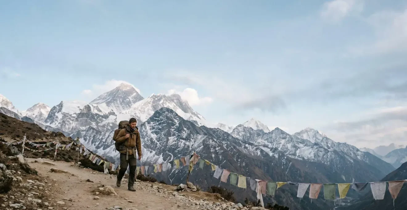 A lone trekker walking a wide Himalayan trail with prayer flags and snow-capped peaks in the distance, symbolizing the achievable journey to Everest Base Camp