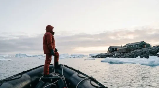 A polar traveler in red expedition gear approaches a cluster of colorful research station buildings on an icy Antarctic bay