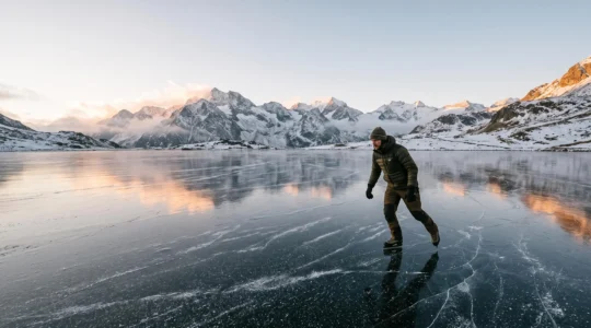 A lone ice skater gliding across a vast, glassy frozen alpine lake surrounded by snow-capped peaks at dawn